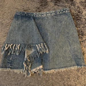 Jeanskjol med fransar - Säljer en cool jeanskjol med omlottkänsla och fransade kanter både fram och bak. Kjolen har en unik design med råa avslut och en liten slits framtill. Perfekt för dig som gillar en edgy stil.