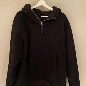 Svart teddyhoodie från Helmut Lang - Mysig svart hoodie från Helmut Lang i teddyfleece med halv dragkedja och stor magficka. Perfekt för kyliga dagar med sin värmande polyester och avslappnade passform. Huvan har snörning för extra komfort.