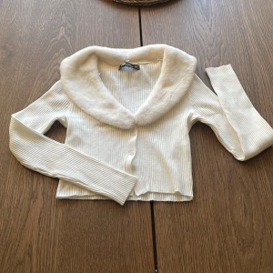 Vit ribbad kofta med fuskpäls-krage från PB Sister Knitwear - Supersöt vit ribbad kofta från PB Sister Knitwear i storlek XS. Tröjan har lång ärm, croppad passform och en fluffig fuskpäls-krage. Stängs med knappar framtill och är perfekt för dig som vill ha något extra till outfiten.