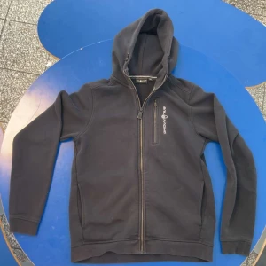 Svart hoodie från Sail Racing med dragkedja - Svart hoodie från Sail Racing med hel dragkedja och bröstficka med dragkedja. Tröjan har huva och logga på bröstet. Tillverkad i bomull och polyester för en mjuk och bekväm känsla.