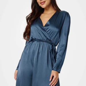 Blå omlottklänning i satin - Snygg långklänning i blå satin med omlottmodell och v-ringning. Klänningen har långa ärmar och knytband i midjan för en smickrande passform. Helt oanvänd med prislapp på.