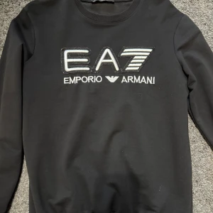 Svart sweatshirt från Emporio Armani EA7 - Säljer en svart långärmad sweatshirt från Emporio Armani EA7. Tröjan har ett stort broderat EA7-logo på bröstet i vitt och silver. Klassisk rund halsringning och mjukt material. Perfekt för en stilren och sportig look.