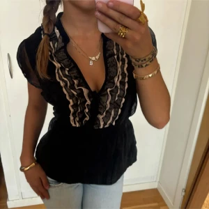 Svart blus med volangdetaljer - Säljer en elegant svart blus med volangdetaljer längs den v-ringade halsen. Blusen har korta ärmar med en lätt transparent look och en smickrande passform. Perfekt för både vardag och fest! 🖤
