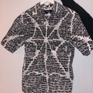 Vit skjorta med svart textmönster från Pacbet - Säljer en vit skjorta från Pacbet med svart, handskriftsliknande textmönster över hela plagget. Skjortan har korta ärmar och klassisk krage. Perfekt för dig som gillar streetwear och unika prints.
