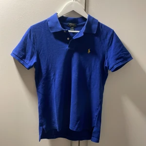 Blå pikétröja från Polo Ralph Lauren - Klassisk blå pikétröja från Polo Ralph Lauren med gul broderad logga på bröstet. Tröjan har krage, korta ärmar och knäppning med två knappar framtill. Perfekt för en stilren och sportig look.