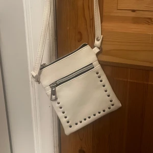 Vit axelväska med nitar - Säljer en stilren vit axelväska med silverfärgade nitar längs kanterna och dragkedja framtill. Väskan har justerbart axelband och är perfekt för att bära det viktigaste. Enkel och snygg design som passar till många outfits.
