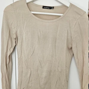 Beige långärmad topp från Boohoo - En enkel och stilren beige långärmad topp från Boohoo. Modellen har rund halsringning och är tillverkad i ett mjukt bomullsliknande material. Perfekt att matcha med jeans eller kjol för en clean look.