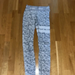 Gråvita mönstrade träningstights från Stronger - Säljer ett par gråvita träningstights från Stronger med marmorerat mönster och två vita ränder på ena benet. Hög midja och elastiskt band med logga upptill. Perfekta för gymmet eller löprundan.