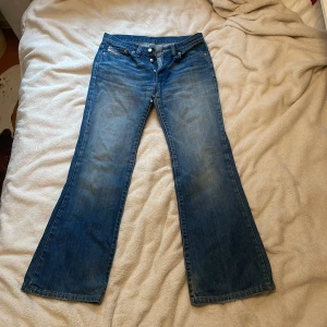 Blå bootcut jeans från Diesel - Säljer ett par klassiska blå bootcut jeans från Diesel med snygg tvätt och detaljerade sömmar. Jeansen har låg midja och en ikonisk Diesel Industry-lapp vid fickan. Perfekta för dig som gillar retrostil.