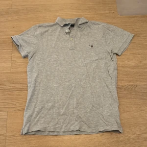 Ljusgrå pikétröja från GANT - Säljer en ljusgrå pikétröja från GANT med klassisk krage och knappar framtill. Tröjan har korta ärmar och en liten broderad GANT-logga på bröstet. Perfekt för en stilren och avslappnad look.