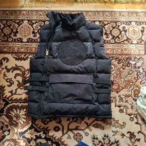 Svart dunväst med broderad logga - Säljer en svart dunväst med stor broderad logga på bröstet och sned dragkedja. Västen har justerbara band i midjan och en stor ficka framtill. Perfekt för lager-på-lager och streetwear-stil.