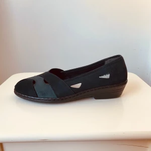 Svarta ballerinaskor från Rieker - Säljer ett par svarta ballerinaskor från Rieker med dekorativa utskärningar och rund tå. Skorna har en låg klack och är tillverkade i läder. Perfekta för dig som vill ha bekväma och stilrena skor till vardags.