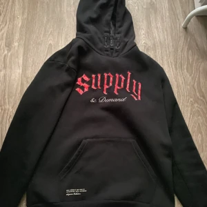 Svart hoodie från Supply & Demand - Svart hoodie med röd och vit text på bröstet där det står 'Supply & demand. Perfekt för en avslappnad stil.