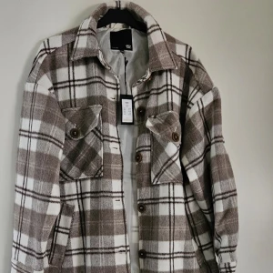 Brun och vit rutig overshirt - Säljer en brun och vit rutig overshirt med stora bröstfickor och knappar framtill. Jackan har klassisk krage och är långärmad, perfekt för lager-på-lager-stil. Mjuk och skön känsla, passar till många outfits.