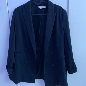  H&M svart kavaj – stilren med modern knäppning - Stilren svart kavaj från H&M! Snygg svart kavaj från H&M med unika detaljer. Modellen har två knappar på båda sidor vilket gör att den kan knäppas lite snett för en trendig look. Tuna vita ränder med långt mellanrum emellan ger ett stilrent intryck. Perfekt för både vardag och fest! Normal passform. 