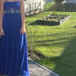 Blå långklänning med strassdetaljer - Säljer en blå långklänning med smala axelband och vackra strassdetaljer i midjan. Klänningen har ett elegant fall och är perfekt för speciella tillfällen. Materialet känns lätt och luftigt.