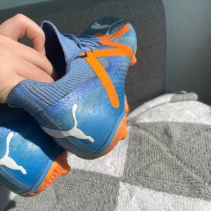 Blå och orange fotbollsskor från Puma - Säljer ett par blå fotbollsskor från Puma med orangea detaljer och vita Puma-loggor. Skorna har snörning och mönstrad sula med dobbar för bra grepp på planen. Perfekta för dig som vill sticka ut på fotbollsplanen!