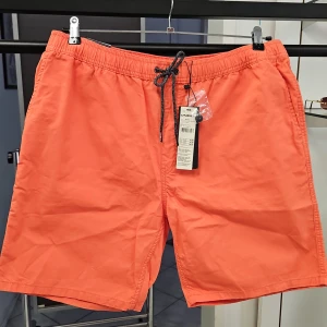 Orange badshorts från O'Neill - Säljer ett par orange badshorts från O'Neill med svart snörning i midjan och elastisk resår. Shortsen har sidofickor och är tillverkade i ett lätt syntetmaterial, perfekta för stranden eller poolen. Vintage effekt. Material/ Bomull.