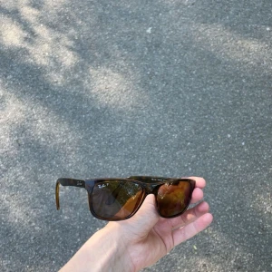 Leopard/Bruna solglasögon från Ray-Ban - Snygga solglasögon från Ray-Ban i brunmelerad plast med klassisk Wayfarer-form. Glasen är mörka och bågarna har en diskret logotyp på sidan. Perfekta för soliga dagar och ger en tidlös look.