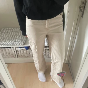 Beiga jeansbyxor med broderad detalj - Säljer ett par beiga jeansbyxor med rak passform och hög midja. Byxorna har en broderad blomma vid benslutet och klassisk femficksdesign. Perfekta för dig som vill ha en unik touch på dina jeans!