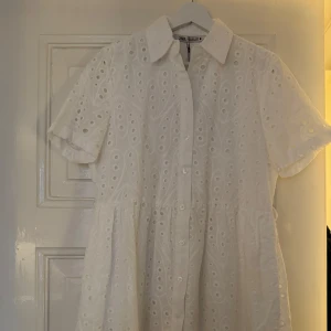 Vit kortärmad klänning med broderade hål och bälte - Söt vit klänning med korta ärmar, knappar framtill och broderade hål i tyget. Klänningen har en klassisk krage och medföljande bälte i midjan för en markerad siluett. Perfekt för varma dagar och somriga tillfällen. 