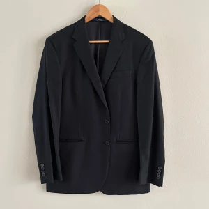 Svart kavaj från Polo Ralph Lauren - Snygg svart kavaj från Polo Ralph Lauren i modellen Custom Fit. Kavajen har två knappar framtill, klassiska slag och fyra knappar vid ärmsluten. En av knappen är trasig men är lätt att byta ut. Perfekt för dig som vill ha en stilren och tidlös look. Enbart använd 1 gång. Nypris 5000kr