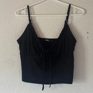 Svart topp från Bik Bok - Säljer en svart topp från Bik Bok med smala axelband och knytning framtill. Toppen har en lätt v-ringning och är perfekt för varma dagar.