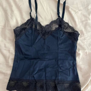 Linne med spets - Säljer ett mörkblått linne med tunna axelband och vackra spetsdetaljer längs halsringning och nederkant. Toppen har en feminin och elegant look, perfekt för att lyfta vilken outfit som helst eller ha som sov eller mys tröja. Köpte den beyond retro men vet ej originellt ”märke”. Gissar på storlek s eller xs, säljer då jag aldrig andvänt den mer än provat 2-3 gånger.