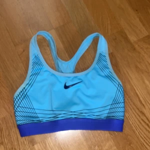 Blå sport-bh från Nike - Säljer en ljusblå sport-bh från Nike med mörkblå detaljer och grafiskt mönster på sidorna. Bred resår nedtill i mörkblått och klassisk racerback-modell. Perfekt för träning och sportiga aktiviteter.