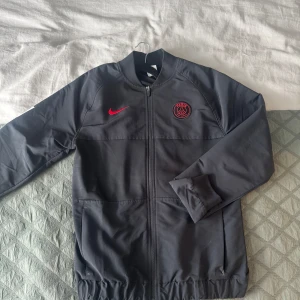 PSG Nike jacka - Säljer en svart PSG träningsjacka från Nike med dragkedja och ribbade muddar. Jackan har PSG-logga och Nike Swoosh i rött framtill samt vit text på ena ärmen. Perfekt för fotbollsträning eller som sportig stil.