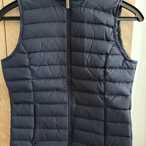 Mörkblå dunväst från Scorpion Bay - Snygg mörkblå dunväst från Scorpion Bay med hög krage och dragkedja framtill. Västen har ett stilrent, quiltat mönster och är perfekt för lager-på-lager. Liten broderad logga på kragen ger en extra detalj.