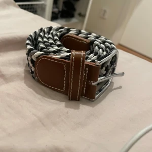 Svart och vit flätat bälte med bruna skinn-detaljer - Säljer ett snyggt flätat bälte i svart och vitt passar perfekt till jeans eller chinos.   Stl 110