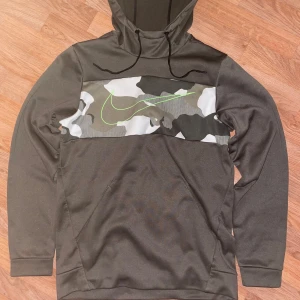 Mörk grön hoodie från Nike med camo-detalj - Säljer en mörk grön Nike hoodie i Dri-Fit material med kamouflagemönster och stor neongrön Nike -logga på bröstet. Tröjan har huva med dragsko och långa ärmar. Perfekt för träning eller chill. Nästan helt oanvänt.