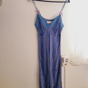 Blå blommig långklänning från Urban Outfitters - Säljer en blå långklänning från Urban Outfitters med blommigt mönster och spetsdetaljer i turkos och rosa vid v-ringningen. Klänningen har smala axelband och en feminin passform. Perfekt för sommarens varma dagar!