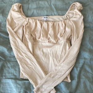 Beige långärmad topp från Bubbleroom - Säljer en beige långärmad topp från Bubbleroom med rynkade detaljer och fyrkantig urringning. Toppen är ribbad och har en feminin passform som passar perfekt till många olika stilar.