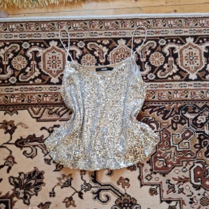 Glittrig silvertopp med paljetter - Säljer en glittrig silvertopp med tunna axelband och v-ringning. Toppen är täckt av paljetter och har en croppad passform, perfekt för att sticka ut på festen. Supersnygg att styla med jeans eller kjol!
