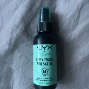 NYX Dewy Finish Setting Spray - NYX Professional Makeup Dewy Finish Setting Spray i svart plastflaska med mintgrön etikett. Ger en daggfräsch finish och håller sminket på plats i upp till 16 timmar. Sprayflaska med genomskinligt mintgrönt lock, innehåller 60 ml.