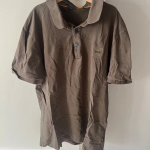 Brun piké t-shirt från BOSS - Säljer en brun piké t-shirt från BOSS i slim fit-modell. T-shirten har korta ärmar, klassisk krage och knappar framtill. Tillverkad i bomull och passar perfekt för en stilren look.