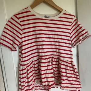 Röd- och vitrandig volangtopp från Zara - Säljer en söt topp från Zara med röda och vita ränder. Toppen har korta ärmar och volang nedtill som ger en lekfull look. Tillverkad i 100% bomull, perfekt för varmare dagar.