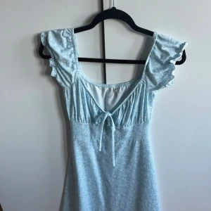 Ljusblå blommig klänning från NLY Trend - Supersöt ljusblå klänning från NLY Trend med småblommigt mönster, volangärmar och knyt framtill. Perfekt för sommarens alla tillfällen. Klänningen har en feminin passform och är tillverkad i ett mjukt material.