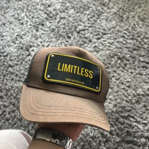 Beige keps från John Hatter & Co med LIMITLESS-tryck - Snygg beige keps från John Hatter & Co med svart och gul patch framtill där det står LIMITLESS. Baksidan är i mesh för extra ventilation och kepsen har justerbar passform. Perfekt accessoar för en cool och avslappnad stil. Kepsen har en liten defekt (repa) på plåten och är solblekt däravpriset. Vid frågor tveka inte att höra av er!😁