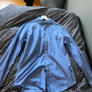 Blå skjorta från Ralph Lauren - Klassisk blå skjorta från Ralph Lauren med button-down krage och broderad logga på bröstet. Skjortan har långa ärmar och vita knappar framtill. Perfekt för en stilren look.