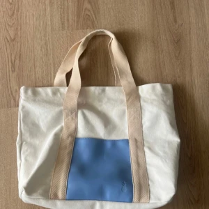 Strandväska - Säljer en stilren vit tygväska med breda beige axelband och en ljusblå ficka framtill. Väskan är rymlig och perfekt för att bära med sig allt man behöver. Enkel och snygg design som passar till många tillfällen. Finns några fläckar (se bild) har inte testat att tvätta den 