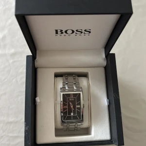 Silverfärgad herrklocka från Hugo Boss - Stilren herrklocka från Hugo Boss med rektangulär svart urtavla och romerska siffror. Klockan har en länkad armlänk i silverfärgat metall och datumvisning. Perfekt accessoar för dig som gillar klassisk design. Original box medföljer. Köpt för cirka 2000kr