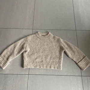 Beige croppad stickad tröja - Säljer en beige croppad stickad tröja med ribbad struktur och lång ärm. Perfekt för lager-på-lager och passar till både jeans och kjol. Hög hals och breda muddar vid ärmslut och nederkant.