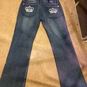 Blå jeansbyxor från Victoria Beckham x Rock & Republic - Säljer ett par blå jeansbyxor från Victoria Beckham x Rock & Republic. Broderade kronor på bakfickorna och tydliga kontrastsömmar. Passa någon som brukar ha storlek s/m