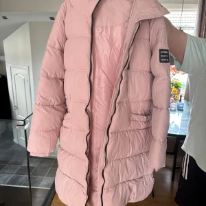 Ljusrosa pufferjacka  - Säljer en ljusrosa pufferjacka med huva och dragkedja. Jackan är lång, har en vadderad design och en patch på ärmen. Perfekt för kyligare dagar och riktigt mysig att bära.