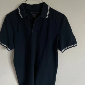 Mörkblå pikétröja från American Eagle - Säljer en mörkblå pikétröja från American Eagle med vita ränder på krage och ärmslut. Klassisk passform och mjukt bomullsmaterial. Perfekt för en stilren och avslappnad look.