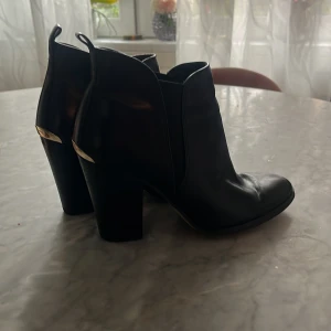 Svarta boots med klack från Michael Kors - Säljer ett par svarta boots från Michael Kors med hög klack och guldfärgade detaljer vid hälen. Skorna är i skinn och har en stilren, elegant design med rund tå. Klacken är 8 cm, använda ett fåtal gånger! ☺️