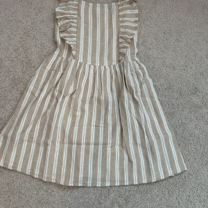 Beige och vitrandig klänning med volang från MNG - Superfin beige klänning med vita ränder från MNG. Klänningen har volangdetaljer längs axelbanden och en smockad rygg för extra komfort. Perfekt för varma dagar med sin luftiga passform och mjuka material.